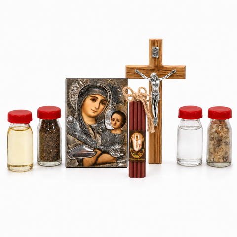 Christian home blessing set arranged on white background with Virgin Mary icon, wooden crucifix, red prayer candles tied with twine, and small glass bottles with liquid, soil and incense