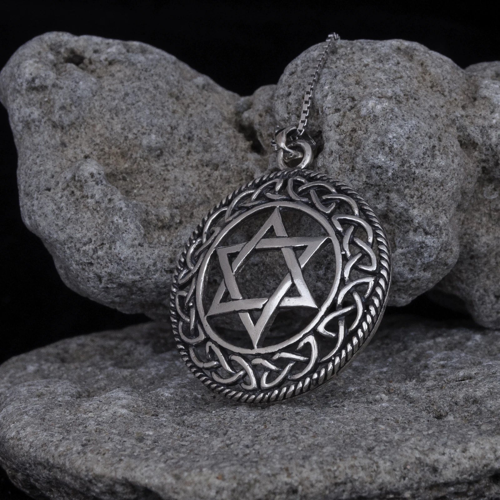 Close-up of a round silver-tone Star of David pendant with dark oxidized recesses, raised Celtic knot border, twisted rope edge, and partial chain, leaning against textured gray rocks on a black background.