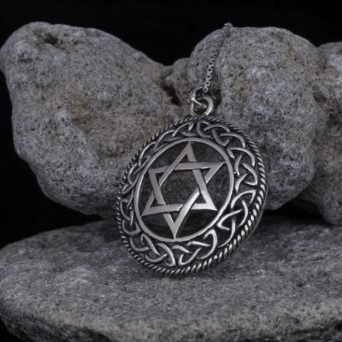 Close-up of a round silver-tone Star of David pendant with dark oxidized recesses, raised Celtic knot border, twisted rope edge, and partial chain, leaning against textured gray rocks on a black background.