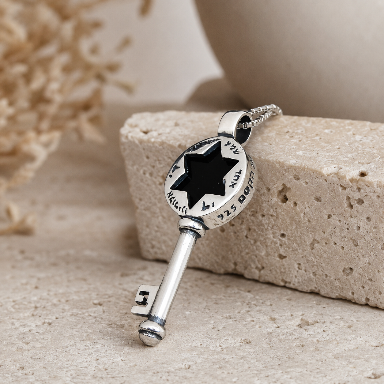 Silver-tone key pendant necklace with black six-point star cutout, Hebrew-style markings around the round top, polished metal finish, angled view on beige stone background
