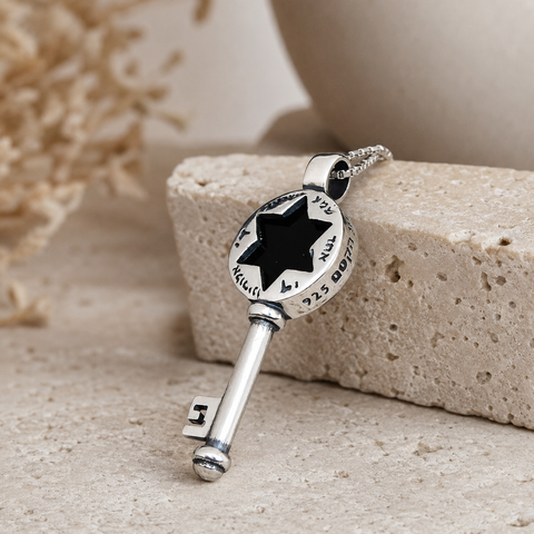 Silver-tone key pendant necklace with black six-point star cutout, Hebrew-style markings around the round top, polished metal finish, angled view on beige stone background